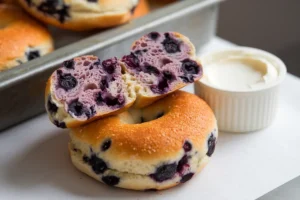 A blueberry bagel is sliced in half and stacked on top of a whole bagel, with cream cheese in a small cup beside them.