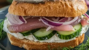 A sandwich with pickled herring, cucumber, red onion, and cream cheese sits on a plate.