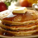 A stack of three pancakes topped with melting butter and syrup sits on a plate.